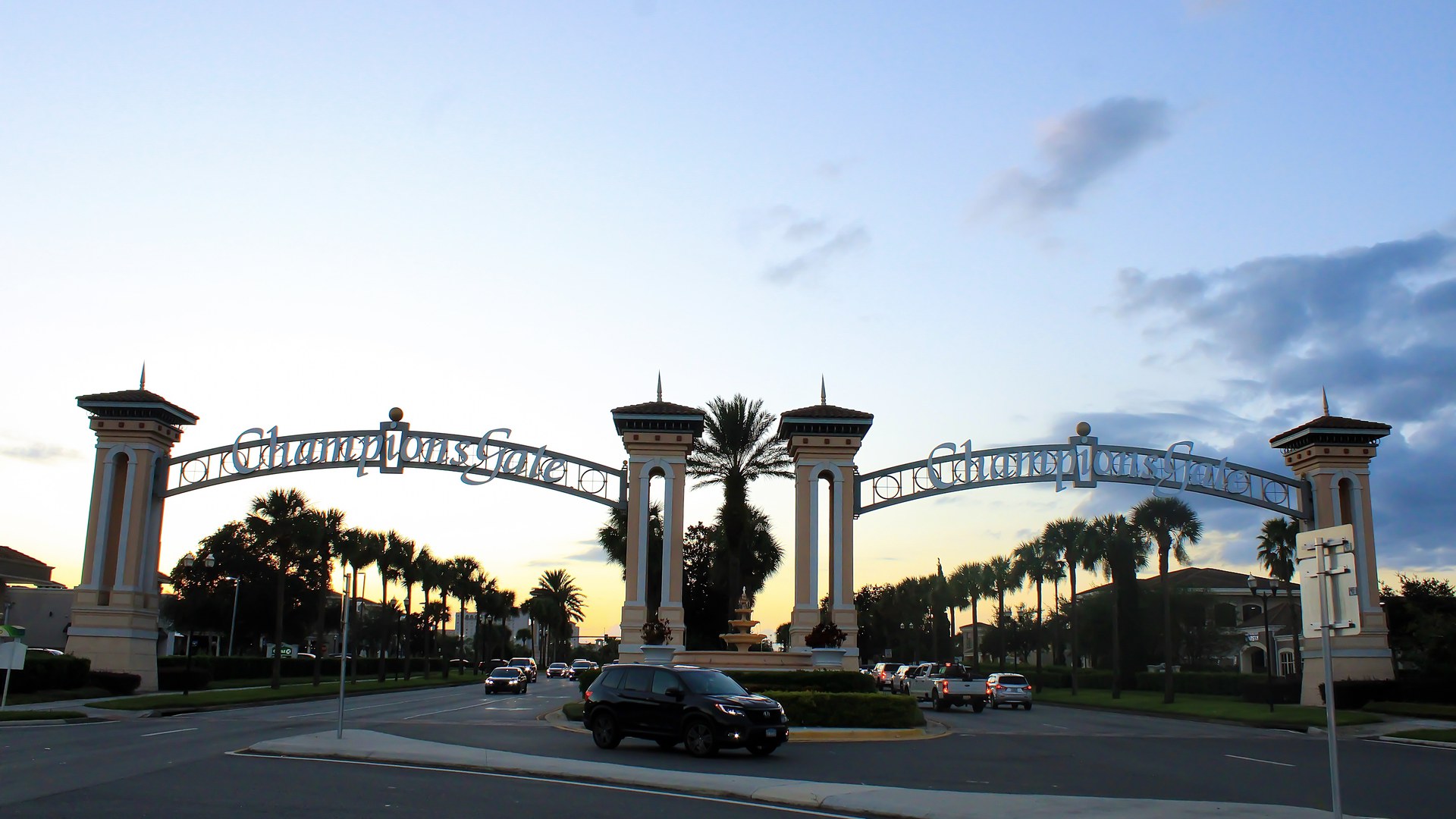 Arcos de entrada de ChampionsGate, comunidade com resort e campo de golfe na região de Orlando, com o nome da comunidade nos arcos, palmeiras e carros na rotatória ao entardecer