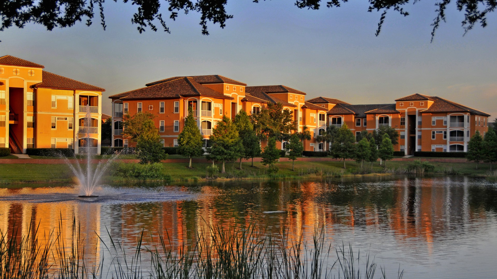 Prédios de apartamentos de três andares, com telhado de telha e fachada laranja, à beira de um lago com chafariz no bairro planejado de MetroWest, em Orlando
