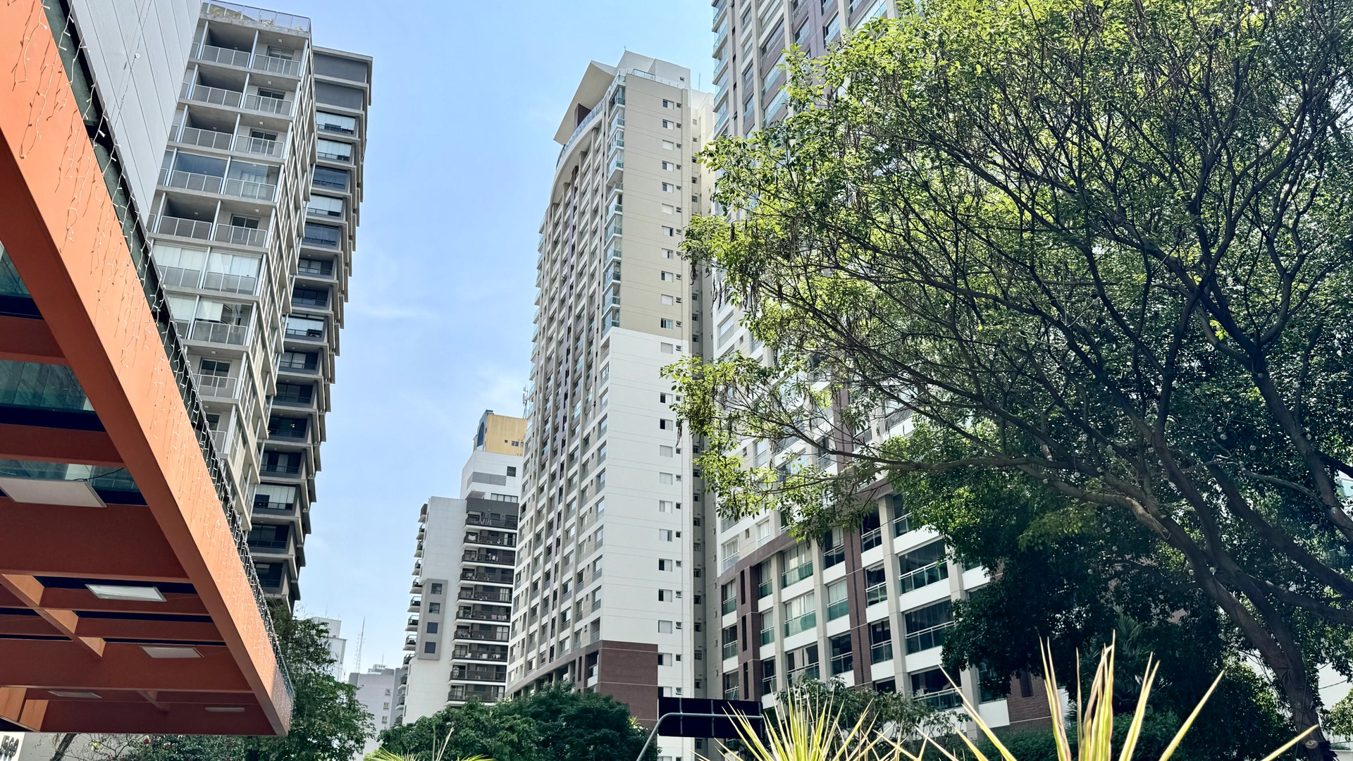 Prédios residenciais altos, com varandas envidraçadas, vistos de baixo numa rua arborizada do Baixo Augusta, em São Paulo, sob céu azul