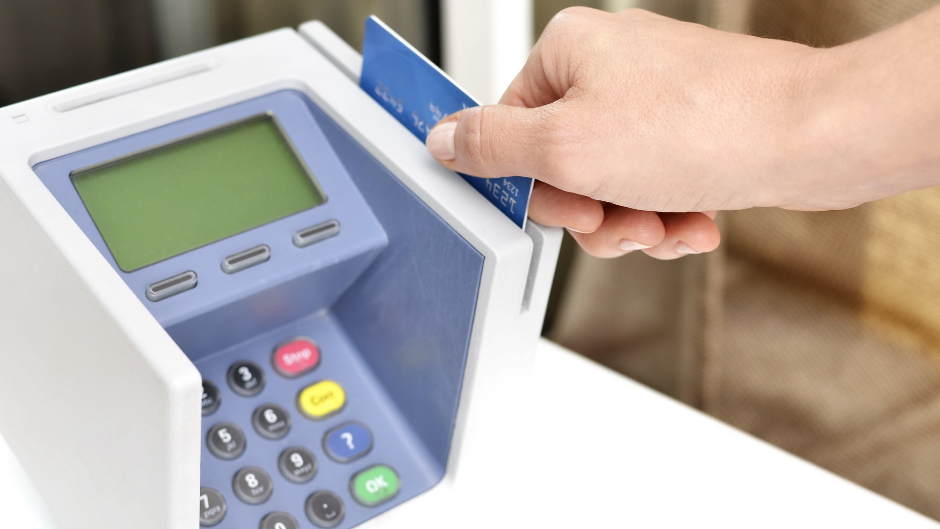 Mão passando um cartão de crédito azul numa maquininha de pagamento branca, com visor verde e teclado numérico, sobre um balcão