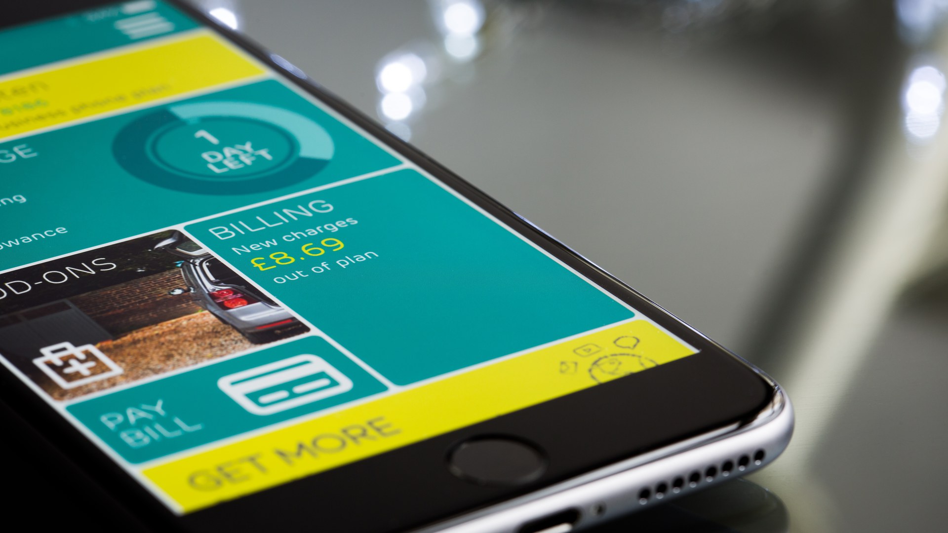 Celular sobre uma mesa de vidro com um aplicativo aberto em blocos verde-água e amarelo, mostrando uma cobrança nova, um botão para pagar a conta com o ícone de um cartão e a faixa «Get more»