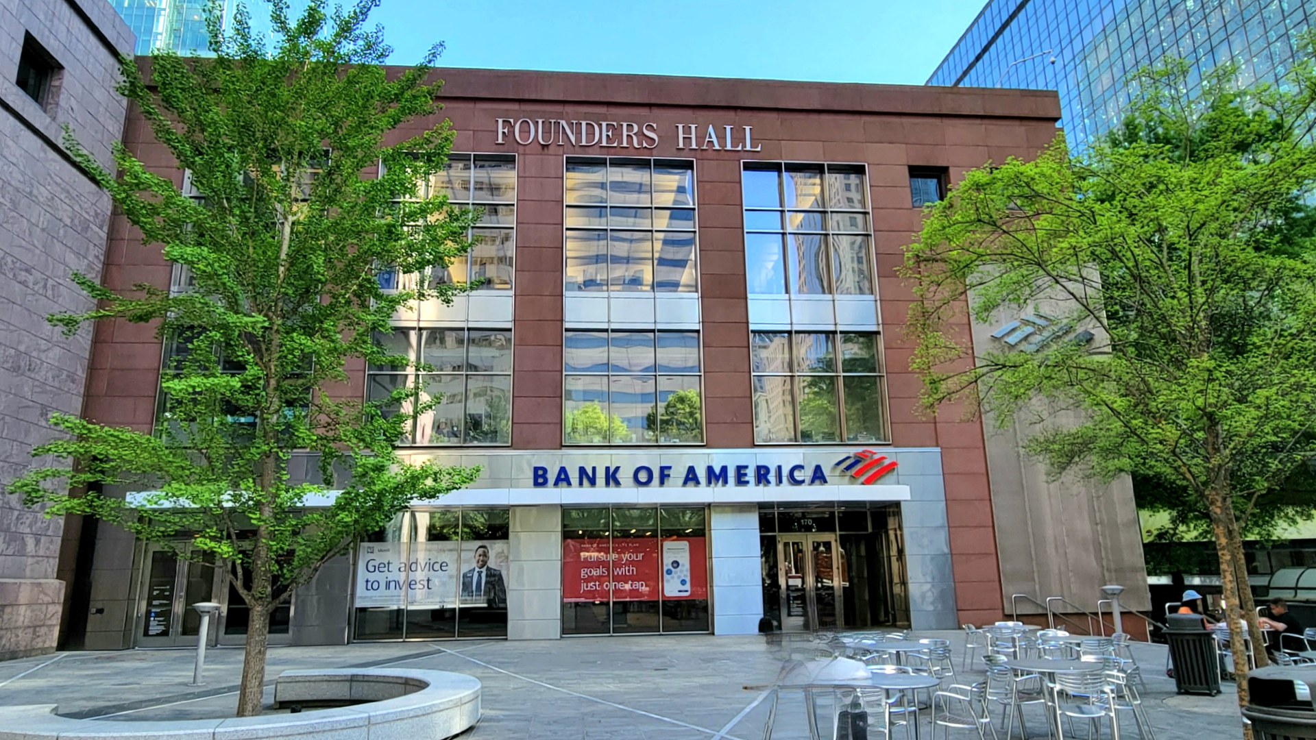 Fachada do Founders Hall, no complexo da sede do Bank of America em Charlotte, na Carolina do Norte, com o letreiro do banco sobre a entrada, árvores e mesas na praça em frente