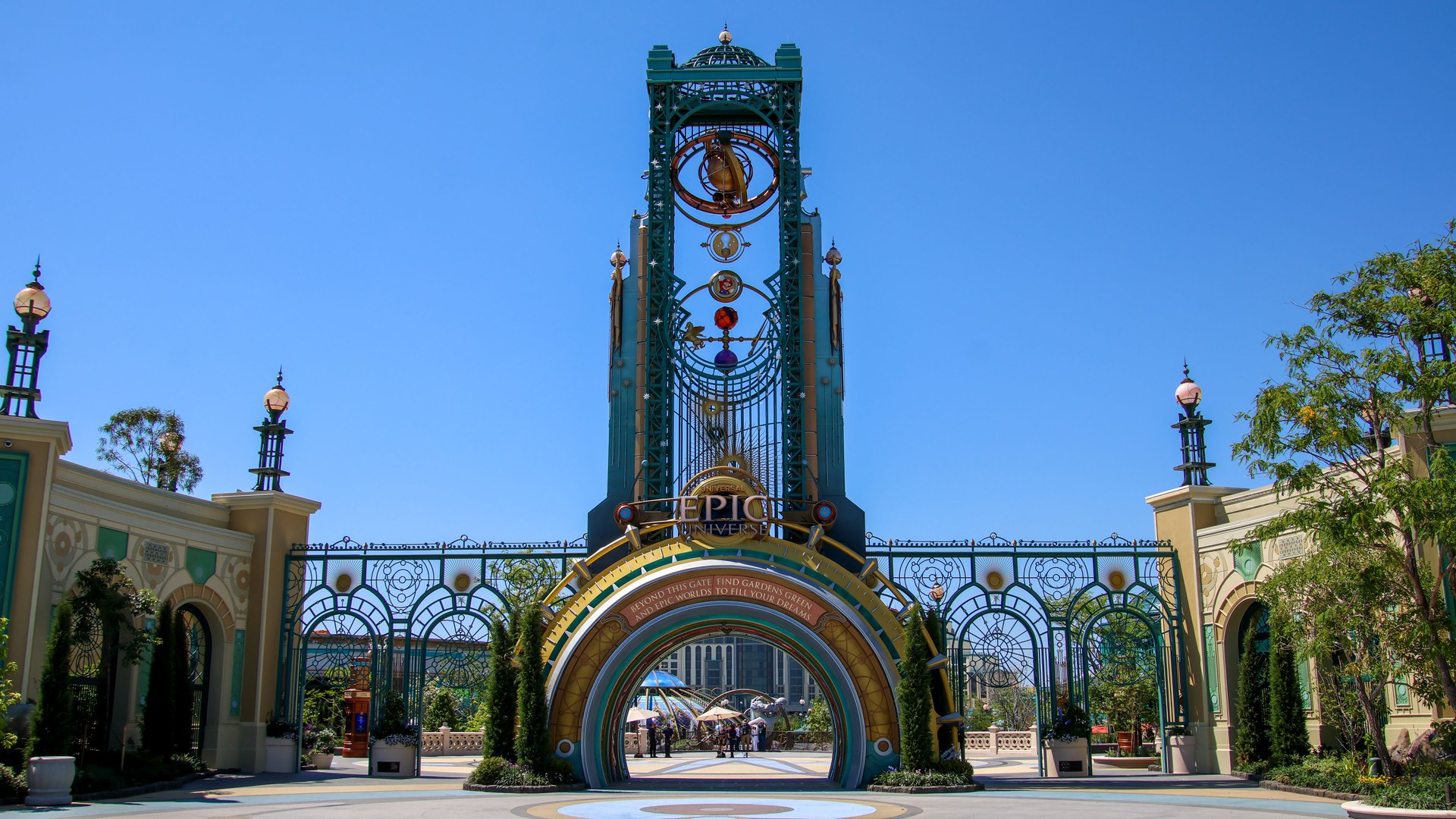 Portão de entrada do Epic Universe, em Orlando, com a torre Chronos verde e dourada sobre um arco com a placa do parque, grades ornamentadas dos lados e céu azul