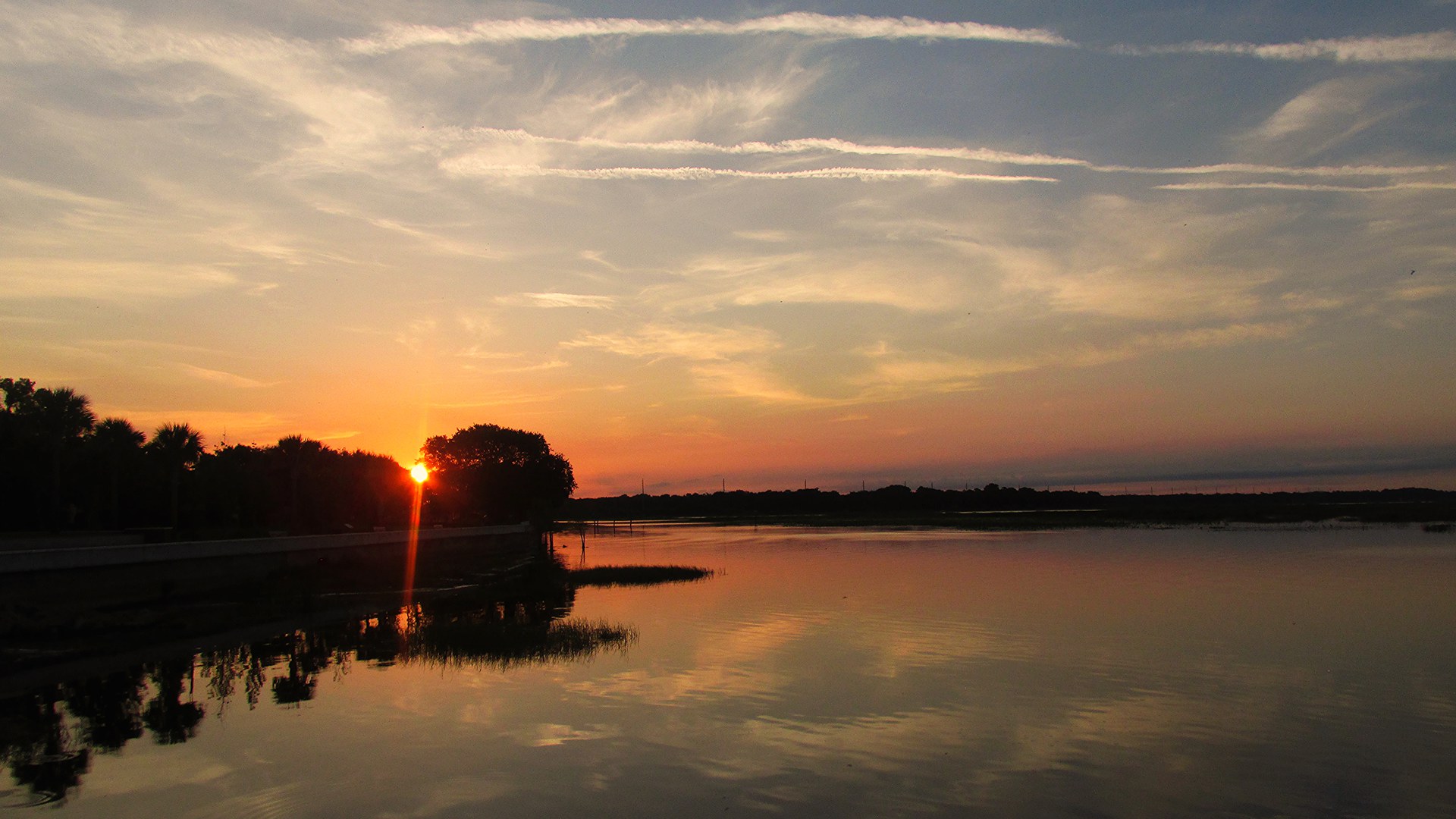 Amanhecer no parque da orla de Kissimmee, à beira do lago Tohopekaliga — o sol nasce entre palmeiras e o céu laranja e azul se reflete na água parada