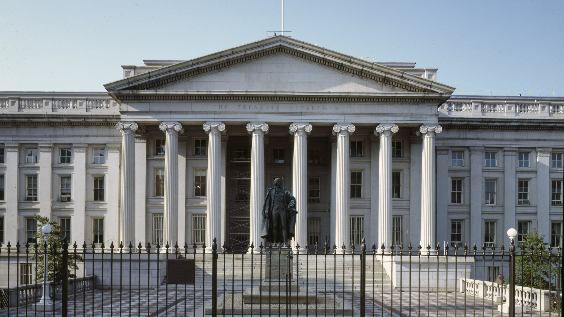 Fachada do prédio do Departamento do Tesouro dos EUA em Washington, com oito colunas, a inscrição "The Treasury Department" no frontão e a estátua de Alexander Hamilton atrás de uma grade de ferro
