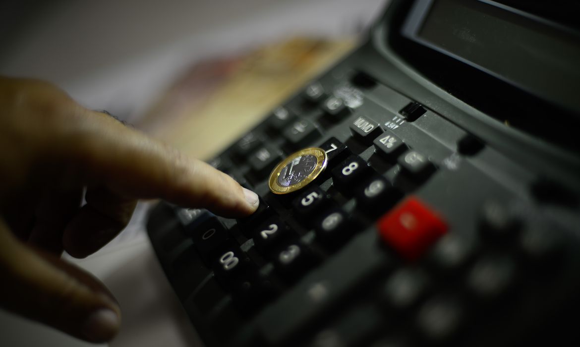 Dedo pressionando a tecla de uma calculadora de mesa preta, com uma moeda de um real apoiada sobre as teclas e cédulas desfocadas ao fundo