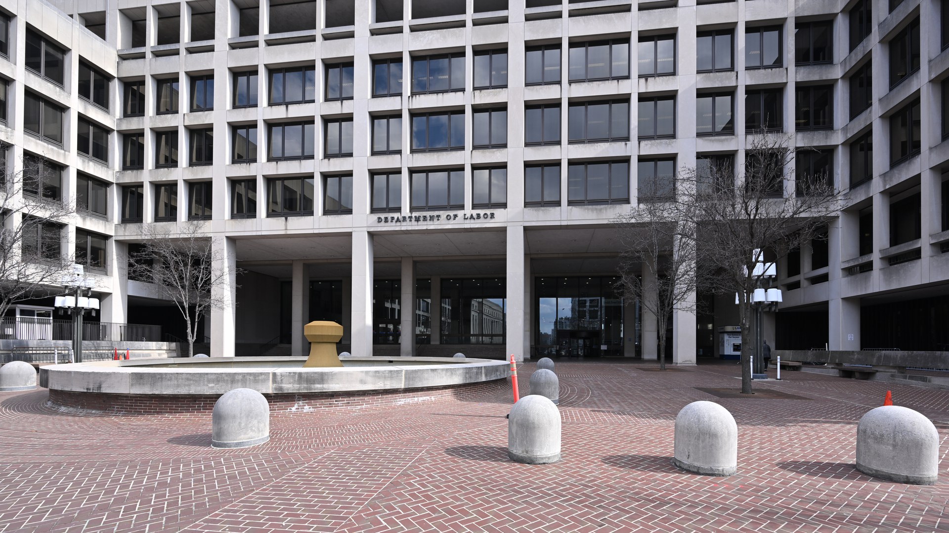 Entrada do Frances Perkins Building, sede do Departamento do Trabalho dos EUA em Washington, com a inscrição "Department of Labor" na fachada, um chafariz e a praça de tijolos à frente