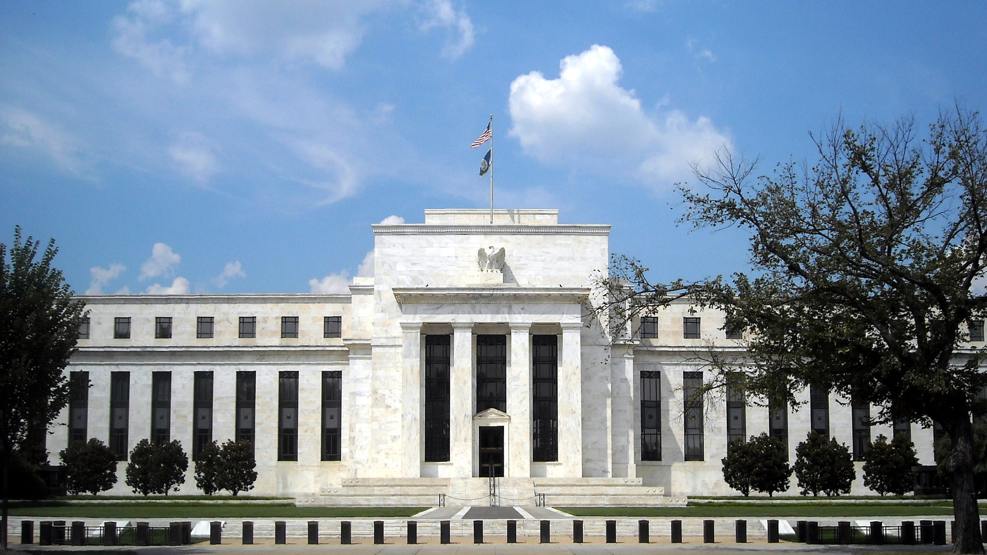 Fachada de mármore branco do Marriner S. Eccles Building, sede do Federal Reserve em Washington, com colunas, uma águia sobre a entrada e as bandeiras dos EUA e do Fed no mastro