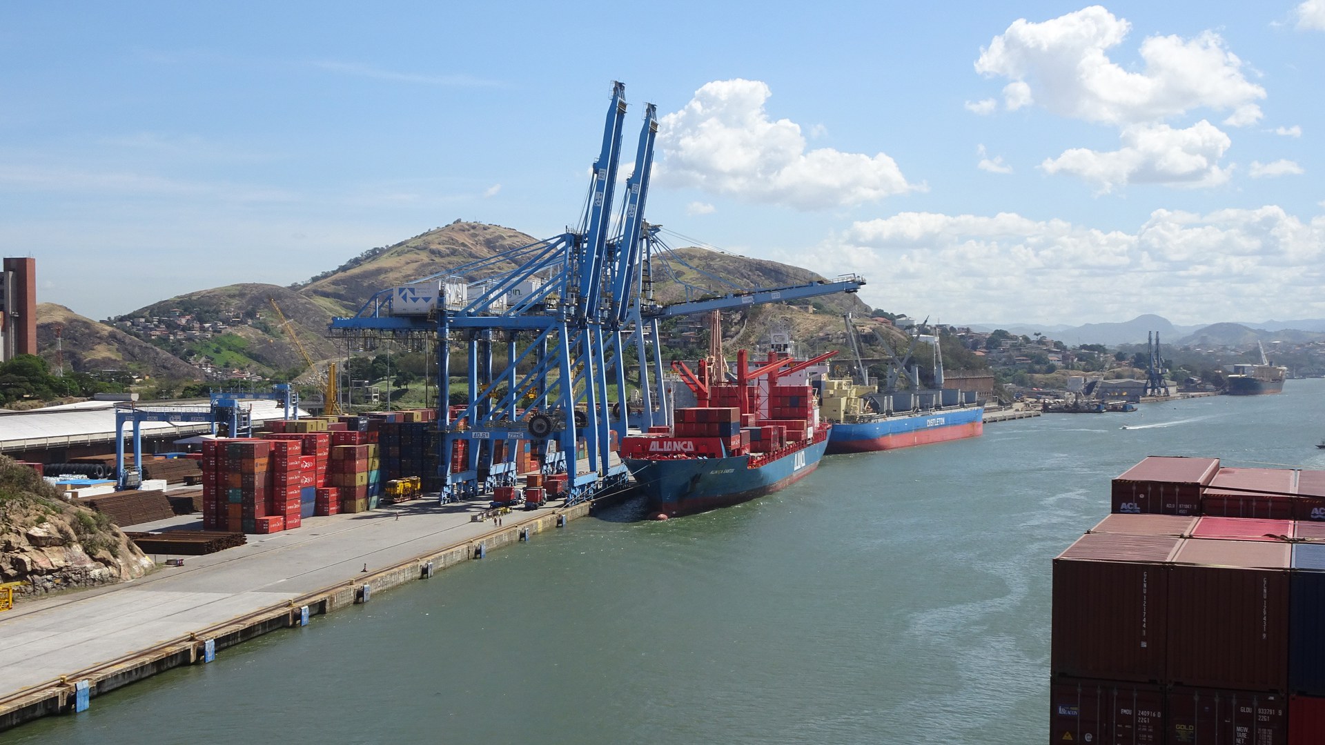Guindastes azuis descarregam um navio porta-contêineres no porto de Vitória (ES), com pilhas de contêineres coloridos no cais e morros ao fundo