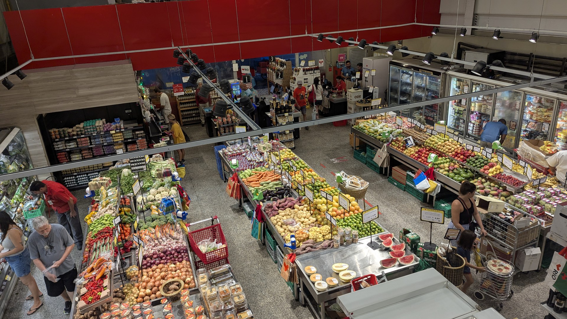 Vista do alto da seção de frutas e verduras de um supermercado no Rio de Janeiro, com bancas de cenoura, batata, cebola, laranja e melancia cortada, plaquinhas de preço e clientes escolhendo produtos