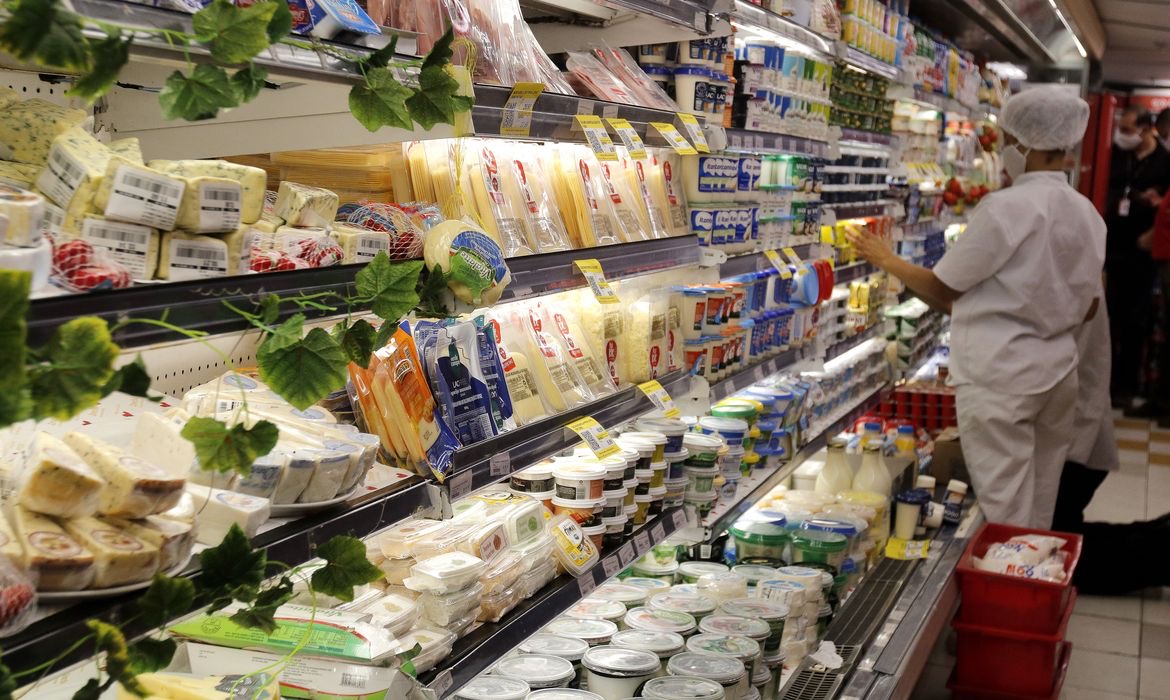 Gôndola refrigerada de frios e laticínios em um supermercado da zona sul do Rio de Janeiro, com uma funcionária de touca e máscara repondo produtos