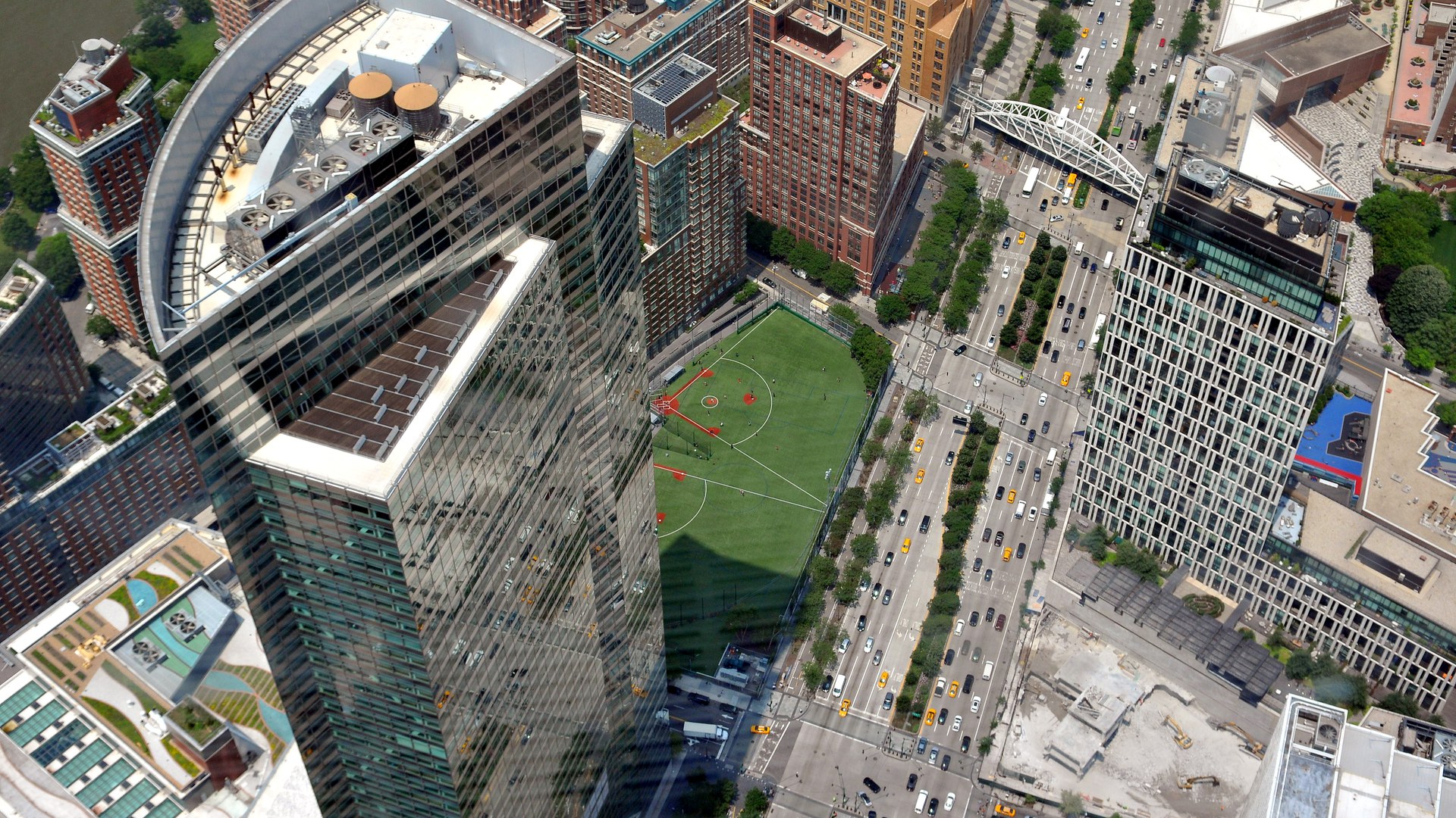 Torre de vidro da sede do Goldman Sachs, em Lower Manhattan, vista do alto, com prédios vizinhos, um campo esportivo e uma avenida movimentada