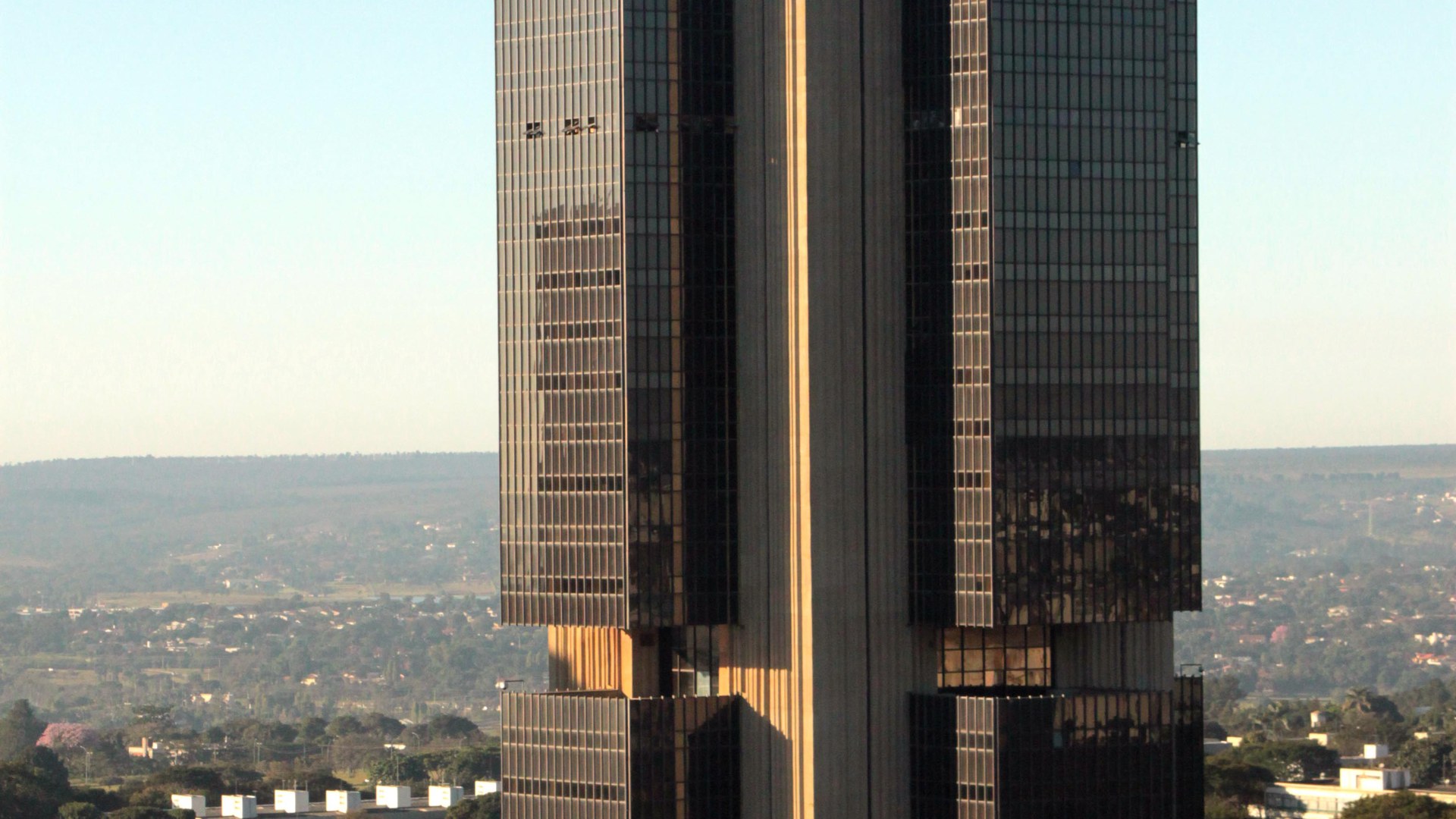 Torre de vidro escuro do edifício-sede do Banco Central em Brasília, com o céu claro e a área verde da cidade ao fundo