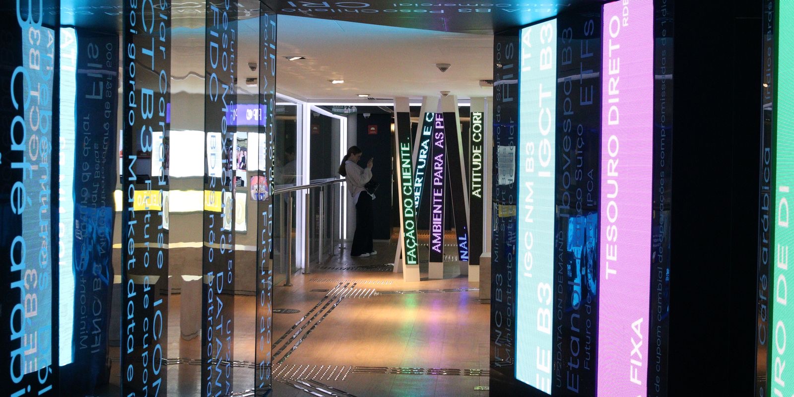 Corredor do Museu da Bolsa do Brasil, no prédio da B3 em São Paulo, com painéis luminosos que exibem nomes de índices e produtos, como Ibovespa e Tesouro Direto