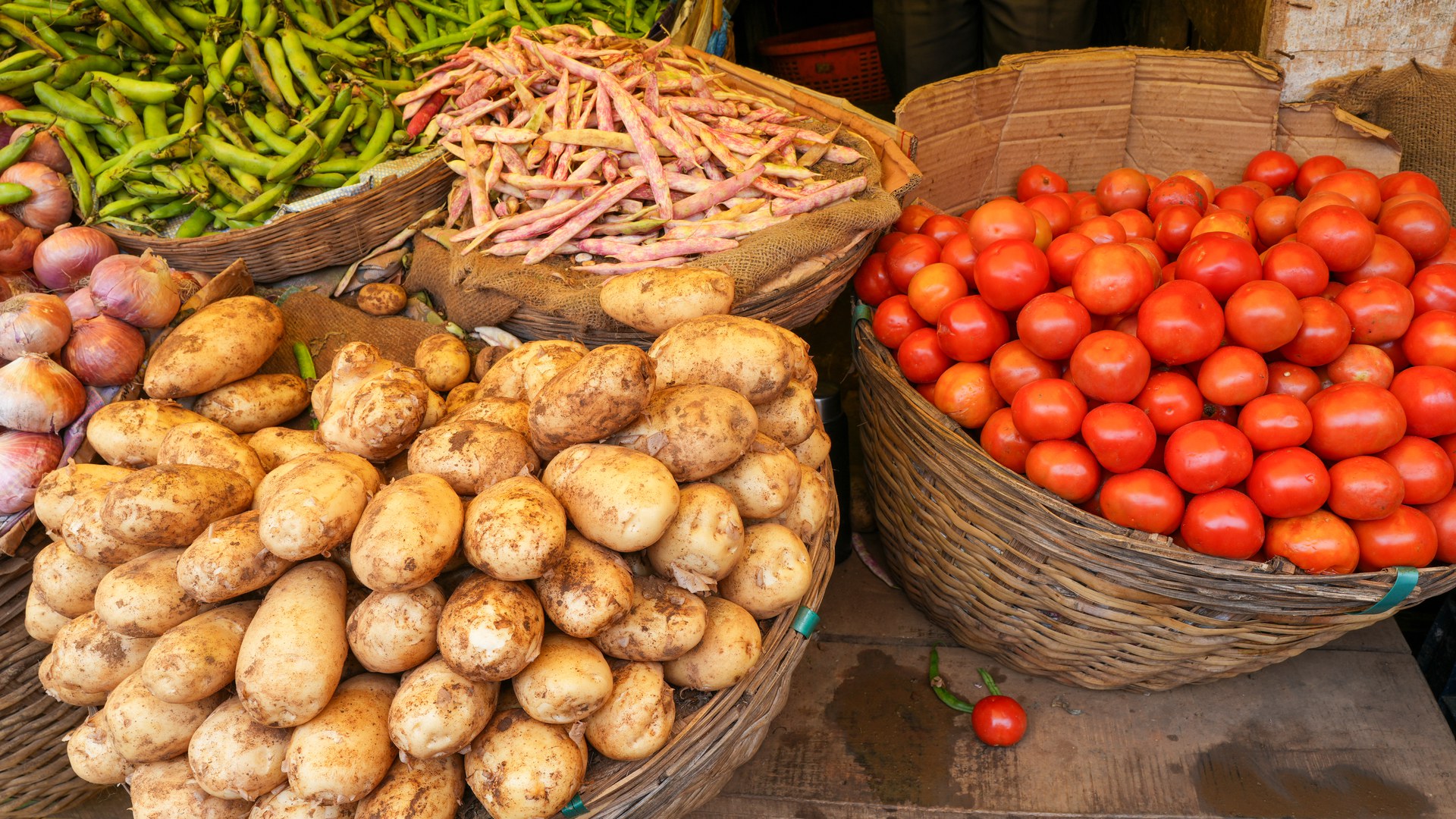 Banca de feira com cestos de vime cheios de batatas em primeiro plano, um cesto de tomates maduros à direita, cebolas roxas à esquerda e vagens ao fundo