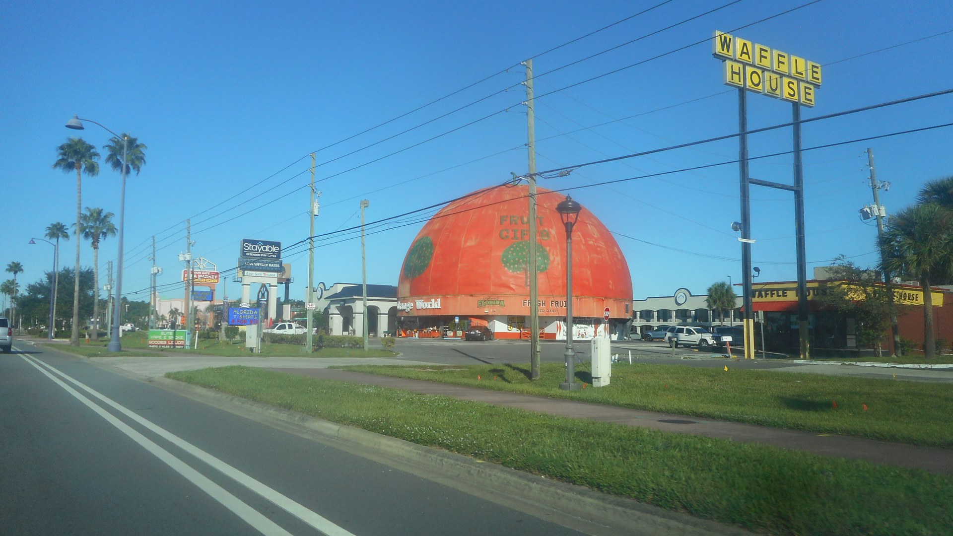 Avenida US 192 perto de Kissimmee sob céu azul, com palmeiras, postes, placas de hotel e lojas e a cúpula laranja da loja Orange World ao fundo