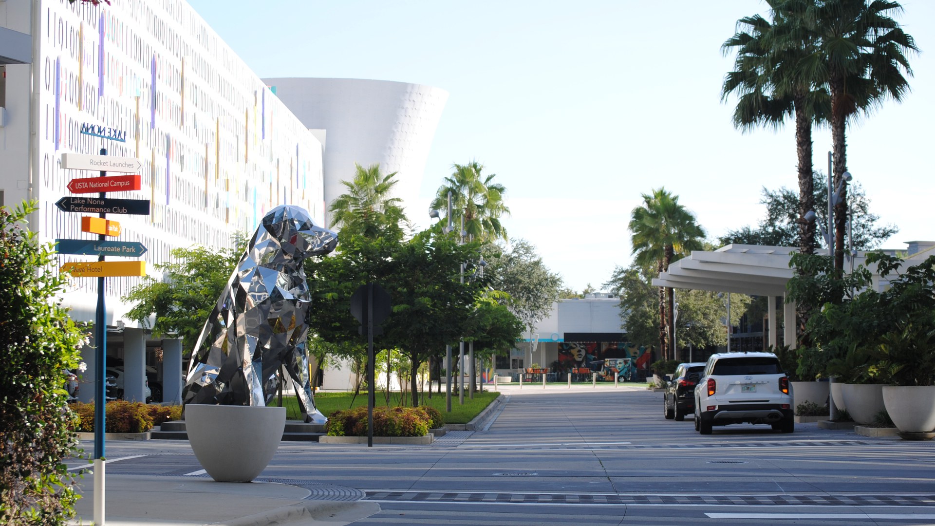 Rua do Lake Nona Town Center, em Orlando, com escultura espelhada de um cachorro, placas de direção para o USTA National Campus e o Laureate Park, palmeiras e carros estacionados