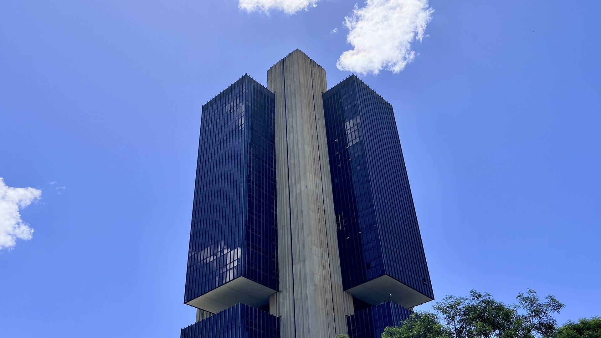 Edifício-sede do Banco Central em Brasília visto de baixo, com duas torres de vidro escuro ligadas por um pilar de concreto claro, sob céu azul com nuvens