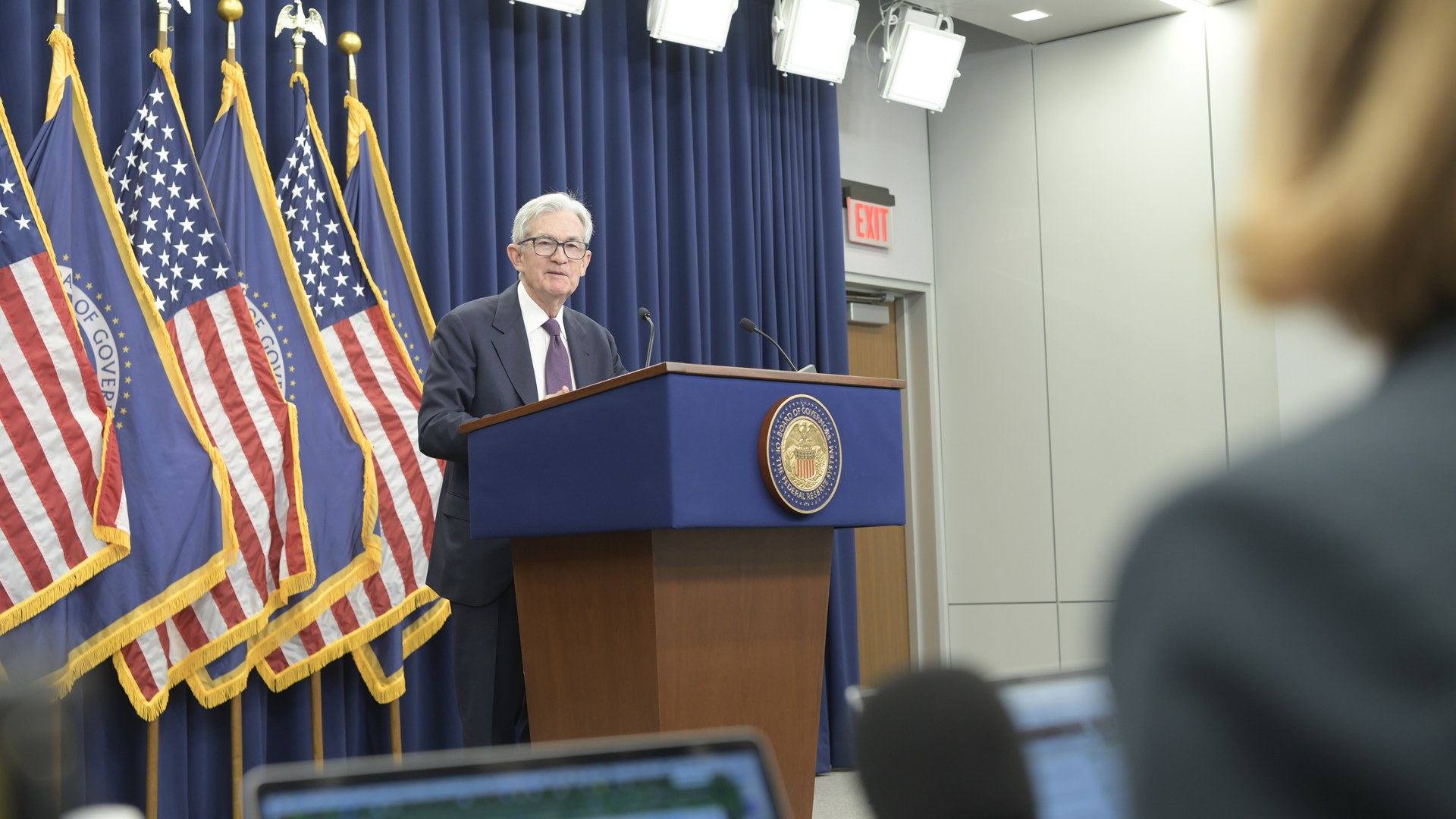 Jerome Powell fala atrás de um púlpito com o selo do Federal Reserve, diante de bandeiras dos EUA e do Fed, em entrevista a jornalistas depois da reunião do FOMC de 29 de abril de 2026; em primeiro plano, notebooks e a cabeça de repórteres desfocados
