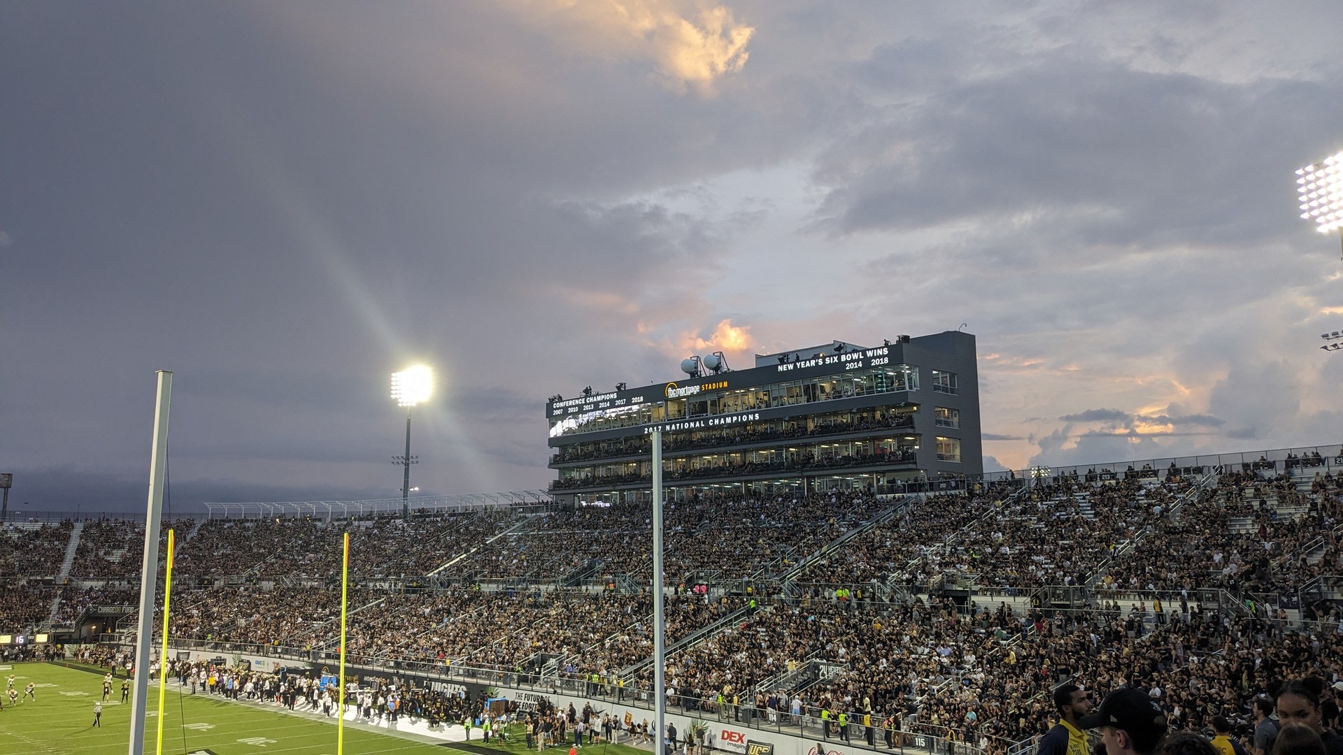 Arquibancadas lotadas do estádio de futebol americano da UCF ao entardecer, com o prédio de camarotes e cabines de imprensa ao centro, refletores acesos e o gramado à esquerda