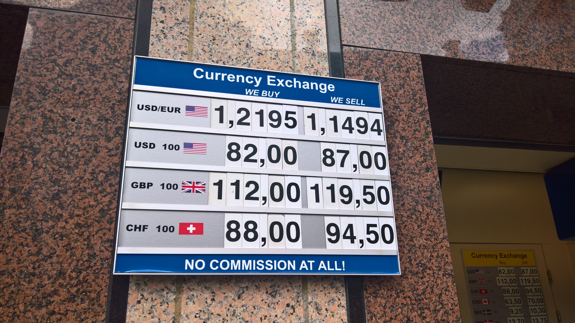 Placa azul e branca de uma casa de câmbio presa a uma parede de granito, com o título «Currency Exchange» e as cotações de compra e venda do dólar, da libra e do franco suíço em números grandes