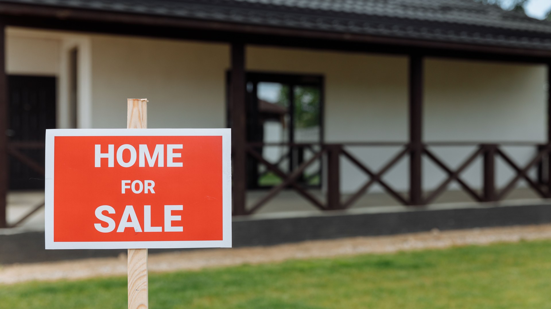 Placa vermelha de «Home for Sale» fincada no gramado diante de uma casa térrea com varanda
