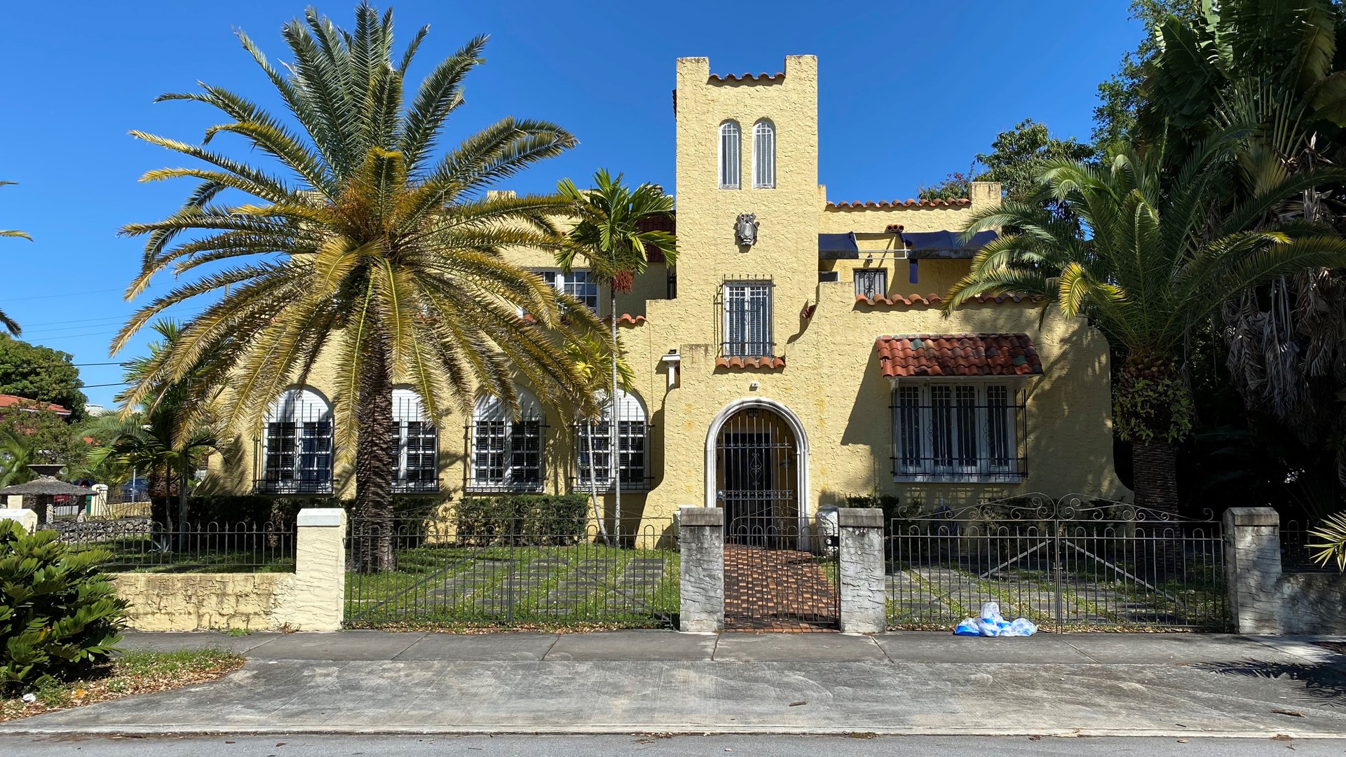 Casa amarela de dois andares em estilo espanhol, com torre central, telhas de barro, janelas com grades e grandes palmeiras atrás de um portão de ferro, numa rua do bairro Shenandoah, em Miami