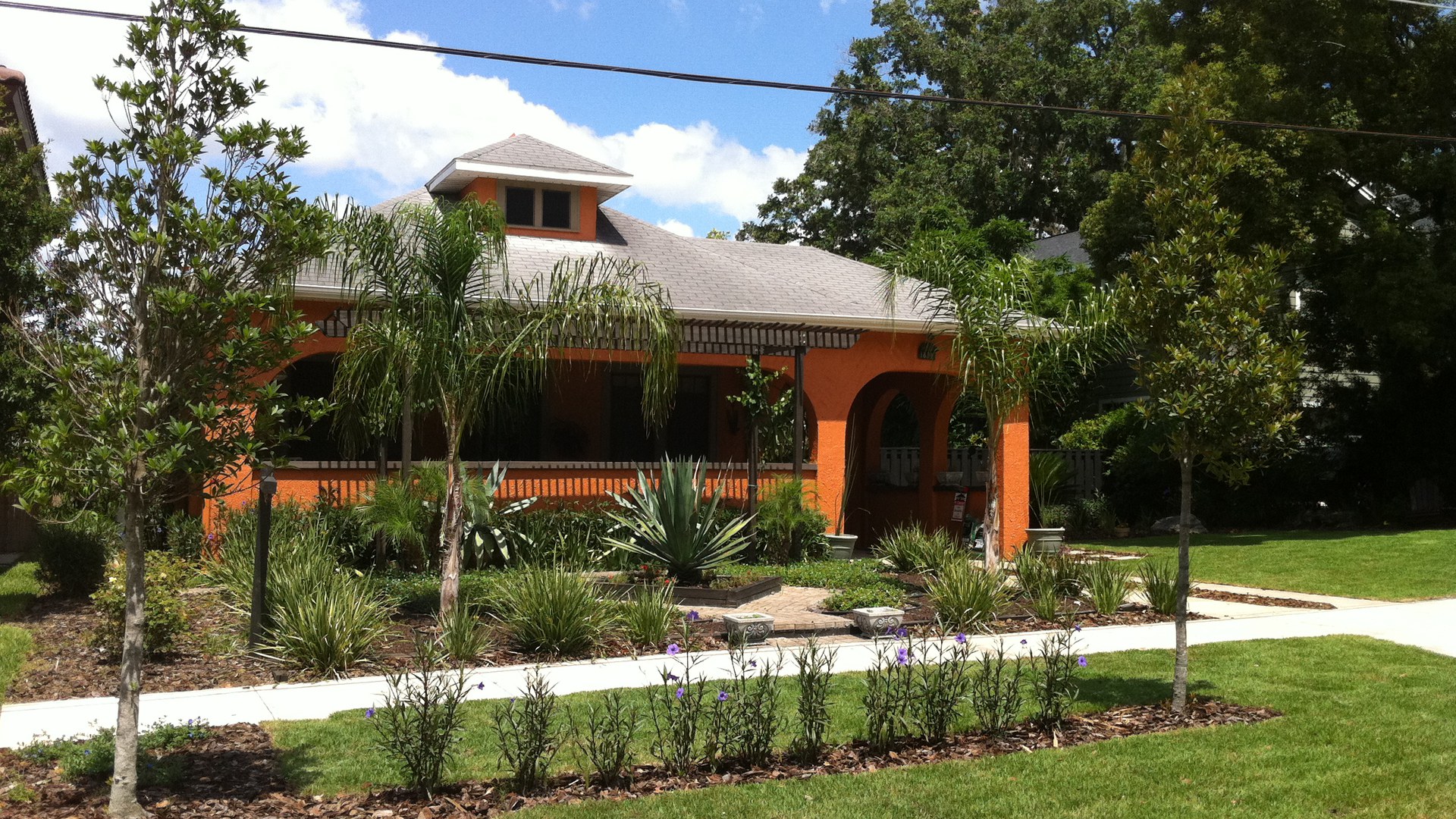 Casa térrea laranja com varanda em arcos e telhado cinza, cercada de palmeiras e jardim tropical, numa rua arborizada de College Park, em Orlando