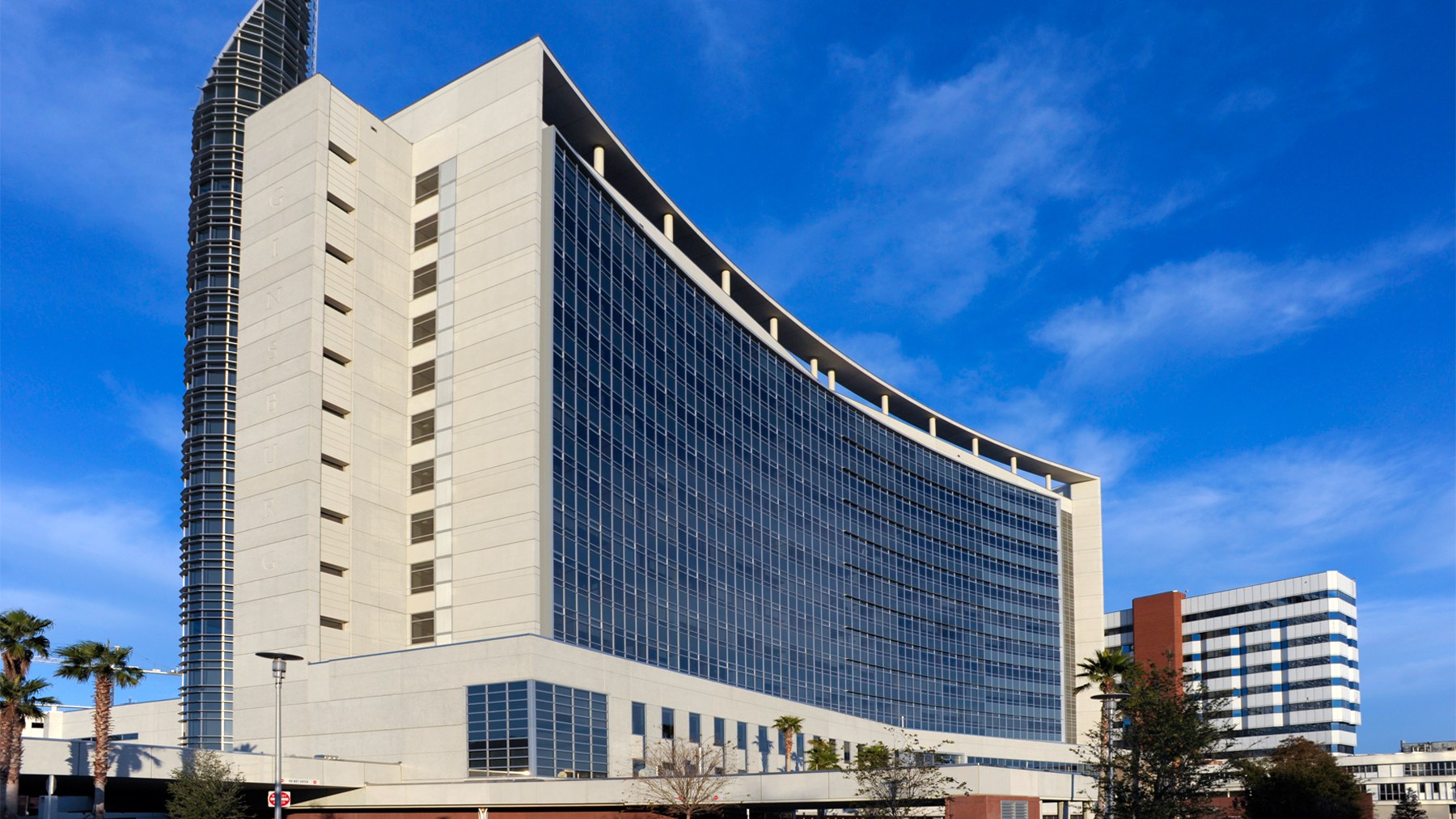 Torre de vidro azul e fachada branca curva do hospital AdventHealth Orlando, com palmeiras na base, sob céu azul