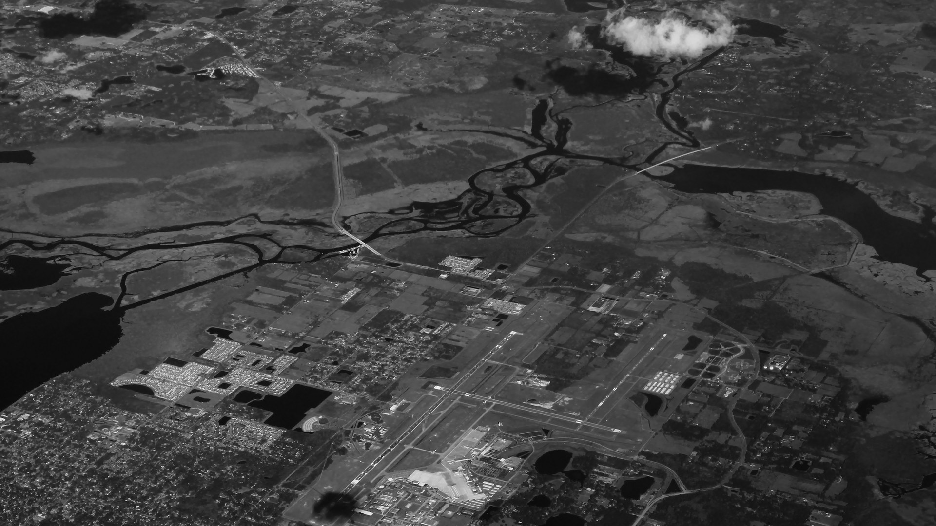 Vista aérea em preto e branco do aeroporto Orlando Sanford, com as pistas cruzadas, os bairros de casas ao lado e áreas alagadas e rios ao fundo