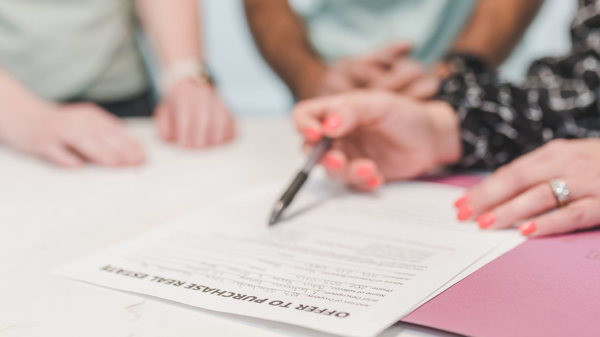 Mão com caneta aponta para um formulário de oferta de compra de imóvel («Offer to Purchase Real Estate») sobre a mesa, com duas pessoas ao fundo.