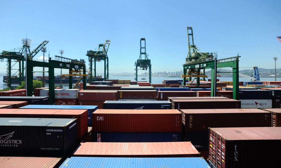 Contêineres empilhados e guindastes no terminal do porto do Rio de Janeiro, com a baía ao fundo