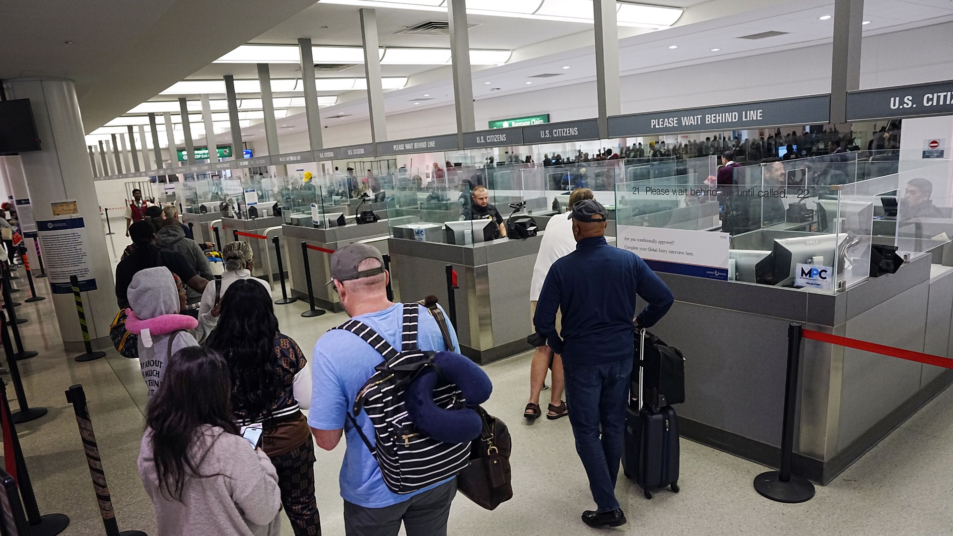 Viajantes vistos de costas, com mochilas e malas, em fila diante das cabines de inspeção de passaportes da alfândega americana (CBP) no aeroporto de Baltimore-Washington, com placas "Please wait behind line"