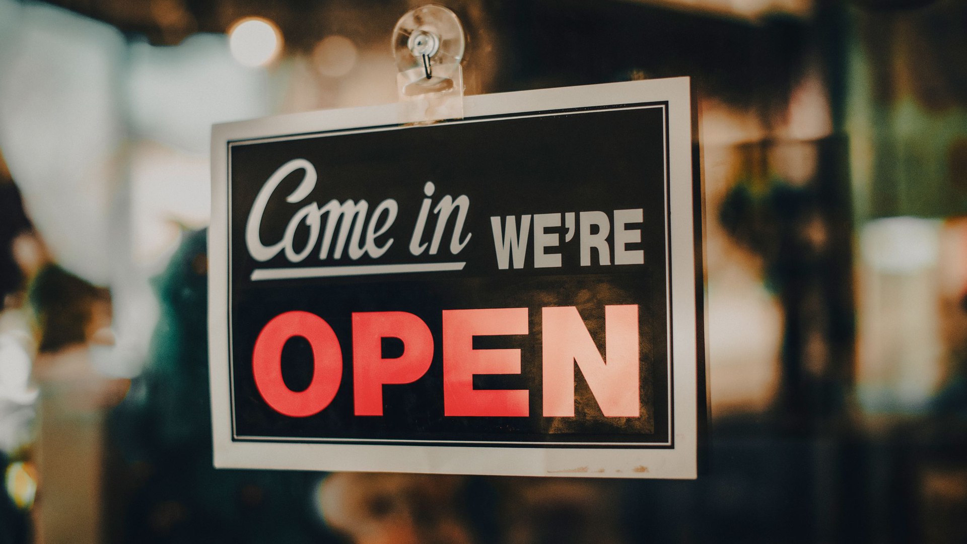 Placa preta com os dizeres «Come in, we're open» e a palavra OPEN em vermelho, pendurada por uma ventosa no vidro da porta de uma loja, com o interior desfocado em luz quente
